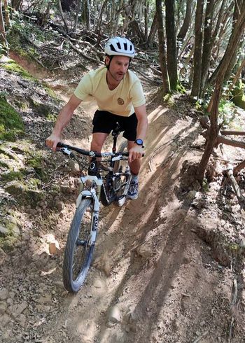 Ein Mann mit weißem Helm und gelbem Hemd fährt auf einem Mountainbike einen schmalen, unbefestigten Weg durch ein Waldgebiet, umgeben von Bäumen und unebenem Gelände.