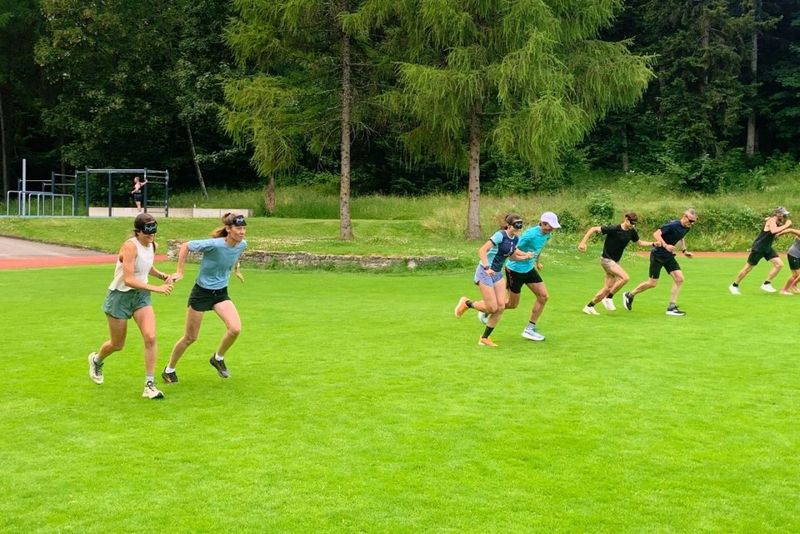 Eine Gruppe von Menschen mit Augenbinden läuft auf einer Wiese, mit Bäumen und einer Bahn im Hintergrund. Einige halten sich an den Händen, andere laufen einzeln.