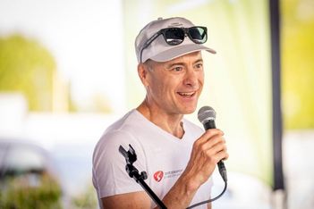 Ein lächelnder Mann in einem weißen Hemd und einer Mütze spricht im Freien in ein Mikrofon. Er hat eine Sonnenbrille auf der Mütze und steht vor einem unscharfen grünen und gelben Hintergrund.