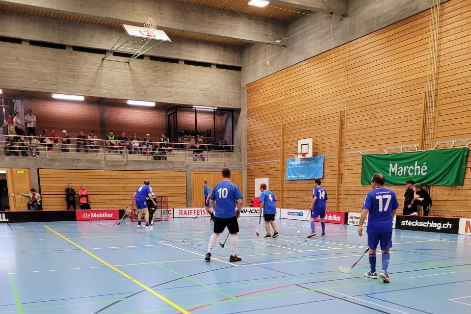 Ein Unihockeyspiel in einer Sporthalle. Spieler in blauen Trikots spielen auf einem hellblauen Spielfeld. Zuschauer beobachten das Spiel von der Tribüne im Hintergrund. Grüne und rote Banner hängen an der Holzwand.
