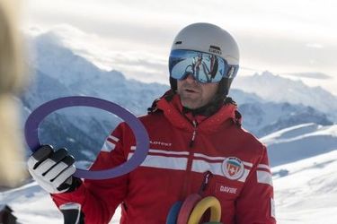 Ein Skilehrer in einem roten Anzug mit bunten Ringen unterhält sich mit einem Skifahrer in schwarzer Ausrüstung auf einem verschneiten Berg mit schneebedeckten Gipfeln im Hintergrund unter einem bewölkten Himmel.