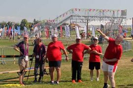 Eine Gruppe von Menschen in roten Hemden sieht zu, wie ein Mann bei einer Sportveranstaltung im Freien einen Ball wirft, mit bunten Fahnen und einer Metallkonstruktion im Hintergrund.