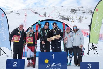Eine Gruppe von sechs Personen posiert auf einem Podium auf einer verschneiten Skipiste, wobei drei Skifahrer auf den obersten Stufen stehen und Auszeichnungen halten. Im Hintergrund sind zwei Fahnen, eine Rennstrecke und Berge zu sehen.