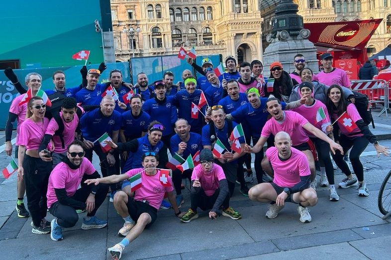 Eine große Gruppe von Menschen, einige in rosa und andere in blauen Hemden, posieren lächelnd mit schweizerischen und italienischen Fahnen auf einem städtischen Platz mit historischen Gebäuden im Hintergrund.