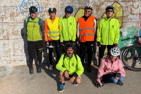 Sieben Radfahrer posieren vor einer mit Graffiti beschmierten Wand. Fünf tragen leuchtend gelbe oder orangefarbene Jacken und Helme, während zwei, darunter ein kniender und ein pinkfarbener, vorne stehen. Die Sonne wirft starke Schatten auf den Boden.