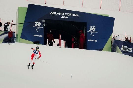 Ein Skifahrer in rot-weißer Kleidung rast am Startpunkt von Milano Cortina 2026 vorbei bergab, während die Mitarbeiter der Veranstaltung in roten Uniformen in der Nähe sind und die Umgebung mit Schnee bedeckt ist.