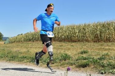 Ein Mann mit Laufprothesen joggt unter blauem Himmel auf einem Kiesweg neben einem Maisfeld. Er trägt ein blaues Hemd, schwarze Shorts und eine schwarze Mütze, die er auf dem Rücken trägt.
