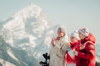 Drei in Winterkleidung gekleidete Personen lächeln und halten Medaillen vor einer verschneiten Bergkulisse. Eine Person hält ein Plüschtier, und alle scheinen in einer kalten Umgebung zu feiern.