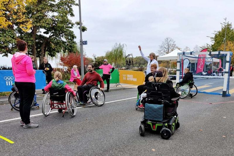 Eine Gruppe von Personen, darunter Kinder und Erwachsene in Rollstühlen, spielt ein Spiel im Freien auf einer gepflasterten Fläche. Zuschauer und Veranstaltungsmitarbeiter in rosa Hemden schauen zu; im Hintergrund sind Herbstbäume und Veranstaltungsbanner zu sehen.