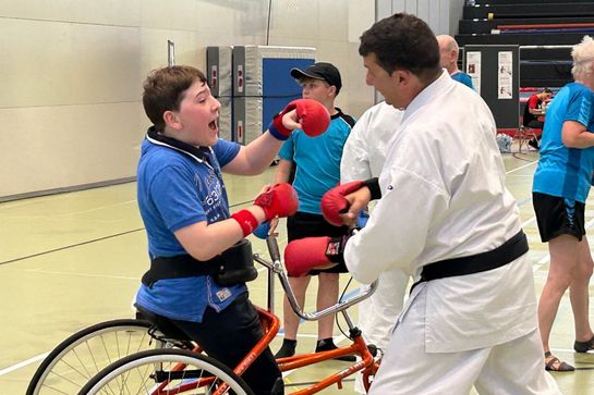 Ein Junge im Rollstuhl mit roten Handschuhen kämpft während einer Indoor-Kampfsportveranstaltung mit einem Mann in weißer Kampfsportuniform. Im Hintergrund sind weitere Teilnehmer und ein Schiedsrichter zu sehen.
