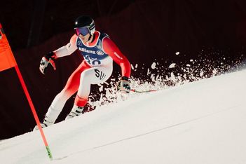 Ein Skifahrer in einem rot-weißen Anzug rast auf einer verschneiten Piste an einer roten Fahne vorbei bergab und wirbelt dabei Schnee auf, während er eine scharfe Kurve fährt. Der Skifahrer trägt eine blaue Startnummer, einen Helm und eine Skibrille.