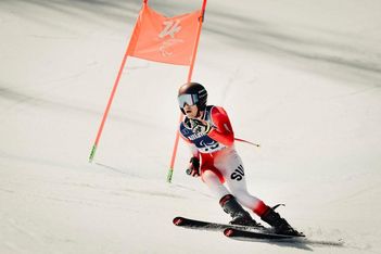 Ein Skifahrer in einem rot-weißen Anzug rast mit Helm und Brille auf einer verschneiten Piste während eines paralympischen Skiwettbewerbs bergab, vorbei an einer orangefarbenen Flagge.