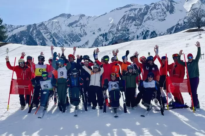 Eine große Gruppe von Skifahrern, von denen einige eine adaptive Skiausrüstung benutzen, posiert auf verschneiten Pisten mit Bergen im Hintergrund. Alle lächeln und recken die Arme in die Höhe und feiern in heller Winterkleidung unter einem strahlend blauen Himmel.