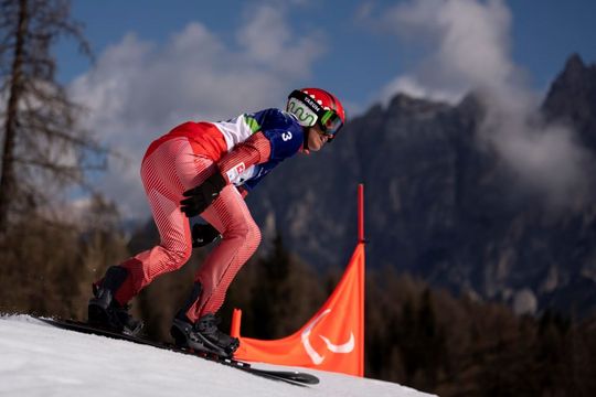 Ein alpiner Skifahrer in rot-weißer Ausrüstung und mit Helm rast an einer orangefarbenen Fahne vorbei einen verschneiten Hang hinunter, mit hohen Bergen und Bäumen im Hintergrund.
