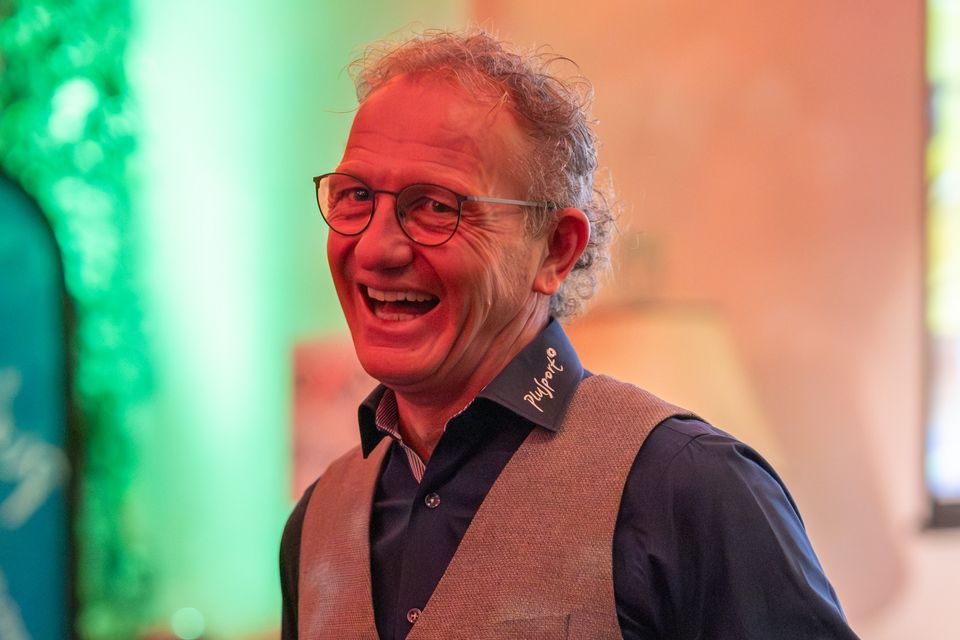 Ein lächelnder Mann mit lockigem Haar und Brille trägt eine graue Weste über einem dunklen Hemd. Der Hintergrund ist mit warmer und grüner Beleuchtung sanft verschwommen.