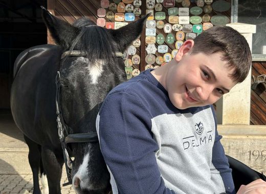 Ein lächelnder Junge lehnt sich in seinem Rollstuhl nach vorn, dicht hinter ihm steht ein schwarzes Pferd. Bunte Plaketten schmücken die Holzwand im Hintergrund.