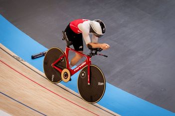 Ein Radfahrer in einem rot-weißen Rennanzug fährt auf einem roten Bahnrad mit schwarzen Scheibenrädern auf einem Indoor-Velodrom und lehnt sich in einer aerodynamischen Position nach vorne.