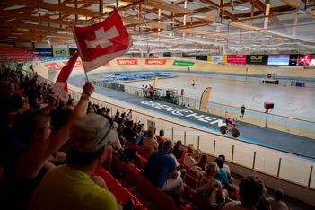Die Zuschauer in einem Velodrom jubeln und schwenken eine Schweizer Fahne, während ein Radfahrer auf der Strecke fährt. Die Arena ist mit Grenchen beschriftet und verfügt über Fahnen, leere Flächen und eine Holzdecke.