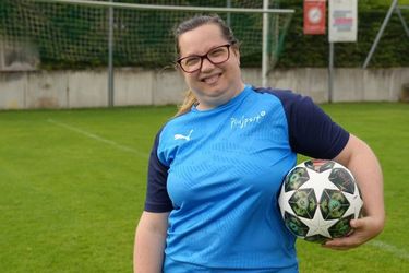 Eine Frau mit Brille und blauem Sporthemd steht auf einem Fußballplatz, lächelt und hält einen Fußball unter dem Arm. Im Hintergrund sind der Torpfosten und der grüne Rasen zu sehen.