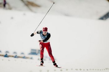 Ein Skilangläufer in roter Hose und roter Mütze fährt auf einer verschneiten Strecke bergauf, wobei er einen Stock benutzt und den anderen in die Luft hebt. Der Skiläufer trägt eine Wettkampf-Latzhose und eine Sonnenbrille. Der Hintergrund ist verschwommener Schnee.