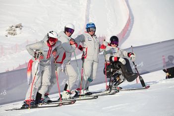 Vier Skifahrer in grauer Kleidung posieren gemeinsam auf einer verschneiten Skipiste, wobei drei auf Skiern stehen und einer einen Sitzski benutzt. Im Hintergrund sind ein rotes Sicherheitsnetz und eine schneebedeckte Landschaft zu sehen.