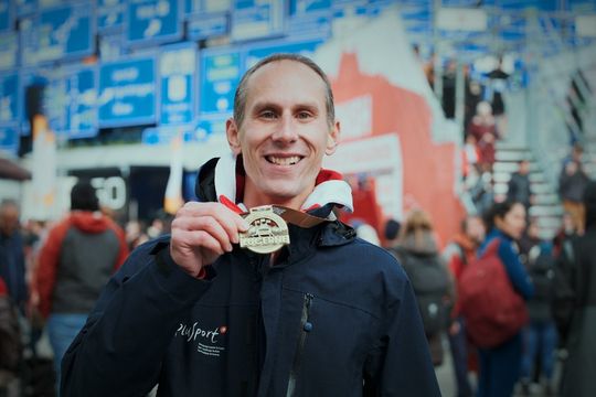 Ein lächelnder Mann in einer Jacke hält vor einer Menschenmenge eine Goldmedaille hoch. Im Hintergrund sind blaue und rote Schilder und Menschen zu sehen, die der Szene eine festliche Atmosphäre verleihen.