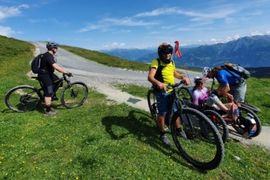 Vier Radfahrer stehen mit ihren Fahrrädern auf einem grasbewachsenen Bergweg unter blauem Himmel. Eine Person trägt ein gelbes Hemd, eine andere ein schwarzes, und zwei weitere, darunter ein Kind, ruhen sich am Wegesrand aus. Im Hintergrund sind Berge zu sehen.