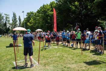 Eine Gruppe von Menschen versammelt sich im Freien auf einer Wiese zu einer Sommerveranstaltung. Die Teilnehmer stehen in einer Reihe hinter einer Seilabsperrung und zwei Personen spielen bei sonnigem Wetter ein Spiel mit einem Ball.