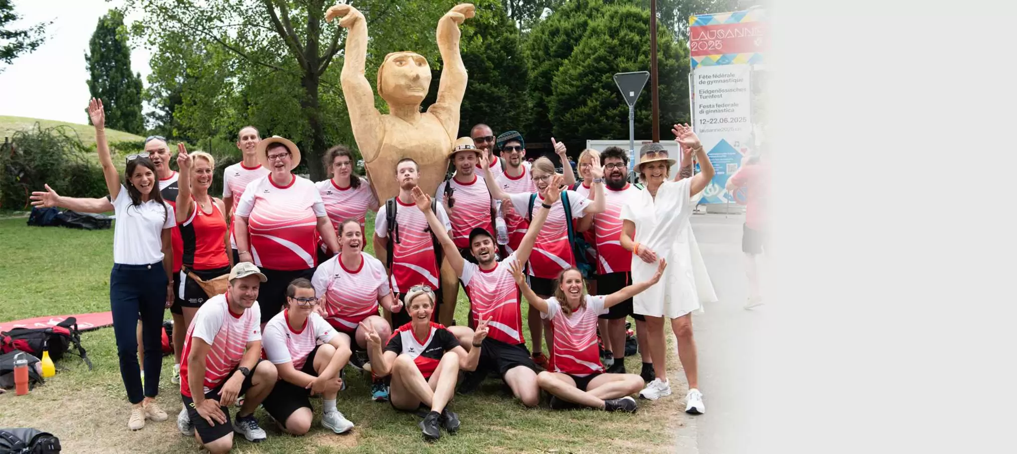 Eine Gruppe von Menschen in passenden rot-weißen Sporthemden posiert und lächelt vor einer großen hölzernen Statue einer Person mit erhobenen Armen, im Freien mit Bäumen und Gras im Hintergrund.