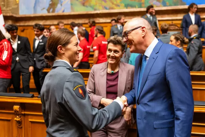 Eine Frau in einer Militäruniform schüttelt einem Mann in einem blauen Anzug die Hand, während eine andere Frau neben ihnen in einem holzgetäfelten Raum voller Menschen lächelt, was auf eine formelle oder offizielle Veranstaltung hindeutet.