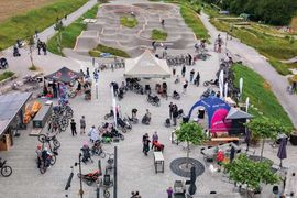 Luftaufnahme eines belebten Fahrradparks mit Menschen, die sich um Zelte, Fahrräder und Rollstuhlfahrräder scharen. Im Hintergrund ist ein gewundener, gepflasterter Pumptrack zu sehen, der von Grün umgeben ist.