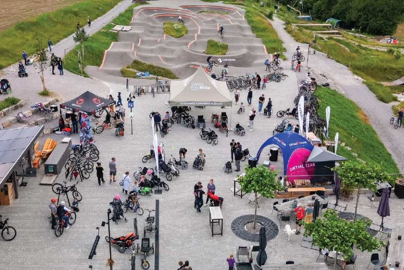 Luftaufnahme eines belebten Fahrradparks mit Menschen, die sich um Zelte, Fahrräder und Rollstuhlfahrräder scharen. Im Hintergrund ist ein gewundener, gepflasterter Pumptrack zu sehen, der von Grün umgeben ist.