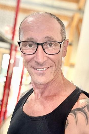 Ein Mann mittleren Alters mit Brille und einem schwarzen Tank-Top lächelt in die Kamera. Er hat eine Tätowierung auf der Schulter und kurzes, schütteres Haar. Der unscharfe Hintergrund deutet auf einen Innenraum hin.