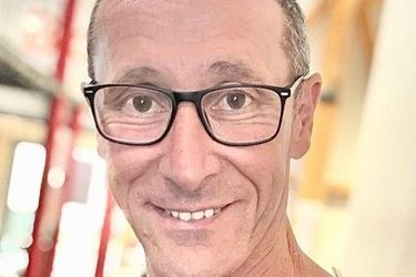 Ein Mann mittleren Alters mit Brille und einem schwarzen Tank-Top lächelt in die Kamera. Er hat eine Tätowierung auf der Schulter und kurzes, schütteres Haar. Der unscharfe Hintergrund deutet auf einen Innenraum hin.