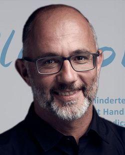 Ein Mann mit Brille und Bart lächelt in die Kamera. Er steht vor einer Kulisse mit den Aufschriften „PluSport“, „Behindertensport Schweiz“, „Sport Handicap Suisse“ und „Sport Handicap Svizzera“ sowie einem Schweizerkreuz als Logo.