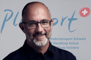 Ein Mann mit Brille und Bart lächelt in die Kamera. Er steht vor einer Kulisse mit den Aufschriften „PluSport“, „Behindertensport Schweiz“, „Sport Handicap Suisse“ und „Sport Handicap Svizzera“ sowie einem Schweizerkreuz als Logo.