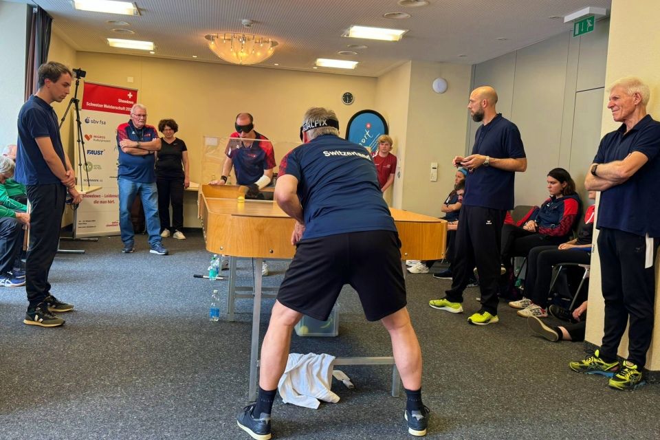 Eine Gruppe von Menschen schaut zwei Männern beim Tischtennisspielen in einem Raum zu. Einige stehen, einige sitzen im Raum. Die Spieler tragen Sportkleidung. Die Atmosphäre ist konzentriert und entspannt. Ein Handtuch und eine Wasserflasche liegen auf dem Boden neben dem Tisch.