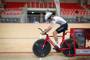 Ein Radfahrer mit weißem Helm und Anzug bereitet sich auf den Start eines Rennens auf einem roten Cervélo-Bahnrad in einem Indoor-Velodrom mit roten Sitzen und einem Holzbelag vor.