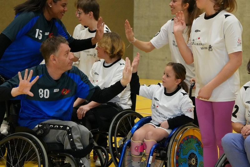 Eine Gruppe von Kindern und Erwachsenen, von denen einige im Rollstuhl sitzen, klatschen sich ab und lächeln, während sie in Sportuniformen in einer Turnhalle gemeinsam feiern.