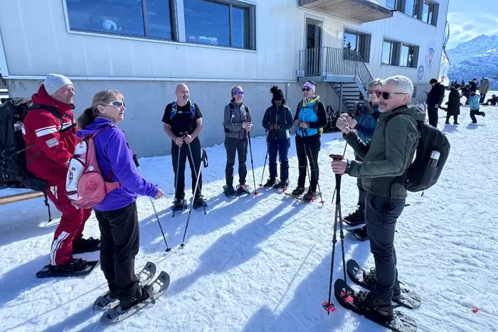 Eine Gruppe von Menschen in Schneeschuhen und Winterkleidung steht im Kreis auf Schnee und hört einem Lehrer in der Nähe eines Gebäudes zu, während im Hintergrund verschneite Berge zu sehen sind.