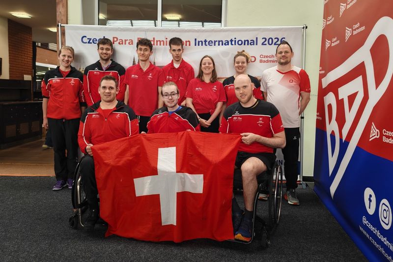 Eine Gruppe von Athletinnen und Athleten in rot-schwarzen Uniformen, darunter zwei Rollstuhlfahrer, posiert in einer Halle mit einer grossen Schweizer Fahne vor einem Banner für das tschechische Para Badminton International 2026.