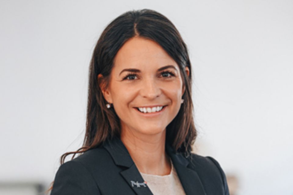 Eine Frau mit langen braunen Haaren, einem dunklen Blazer und einem hellen Oberteil trägt, lächelt und blickt vor einem schlichten, hellen Hintergrund in die Kamera.