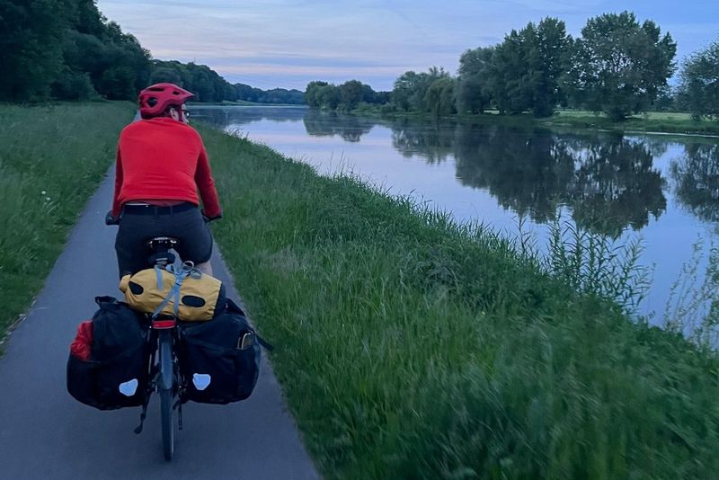 Eine Person mit roter Jacke und Helm fährt mit einem beladenen Tourenfahrrad in der Abenddämmerung einen Uferweg entlang, wobei sich der Himmel rechts in den Bäumen und im ruhigen Wasser spiegelt.