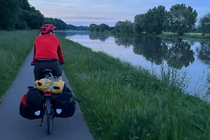 Eine Person mit roter Jacke und Helm fährt mit einem beladenen Tourenfahrrad in der Abenddämmerung einen Uferweg entlang, wobei sich der Himmel rechts in den Bäumen und im ruhigen Wasser spiegelt.