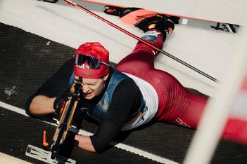 Ein Biathlet, der eine rote Hose und eine rote Mütze trägt, liegt während eines Biathlon-Wettkampfs in Bauchlage auf dem Schnee und zielt mit einem Gewehr, während hinter ihm die Skier zu sehen sind.