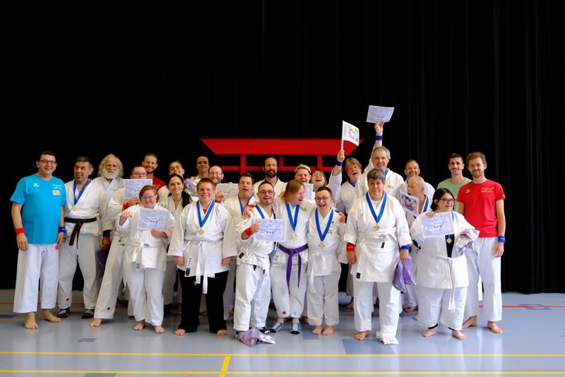 Eine Gruppe von Menschen in Kampfsportuniformen steht in einem Innenraum zusammen, lächelt und hält Urkunden und Medaillen in der Hand. Einige tragen farbige Gürtel, und ein paar Personen in roten und blauen Hemden stehen am Rande der Gruppe. Hinter ihnen befindet sich eine rote Struktur.