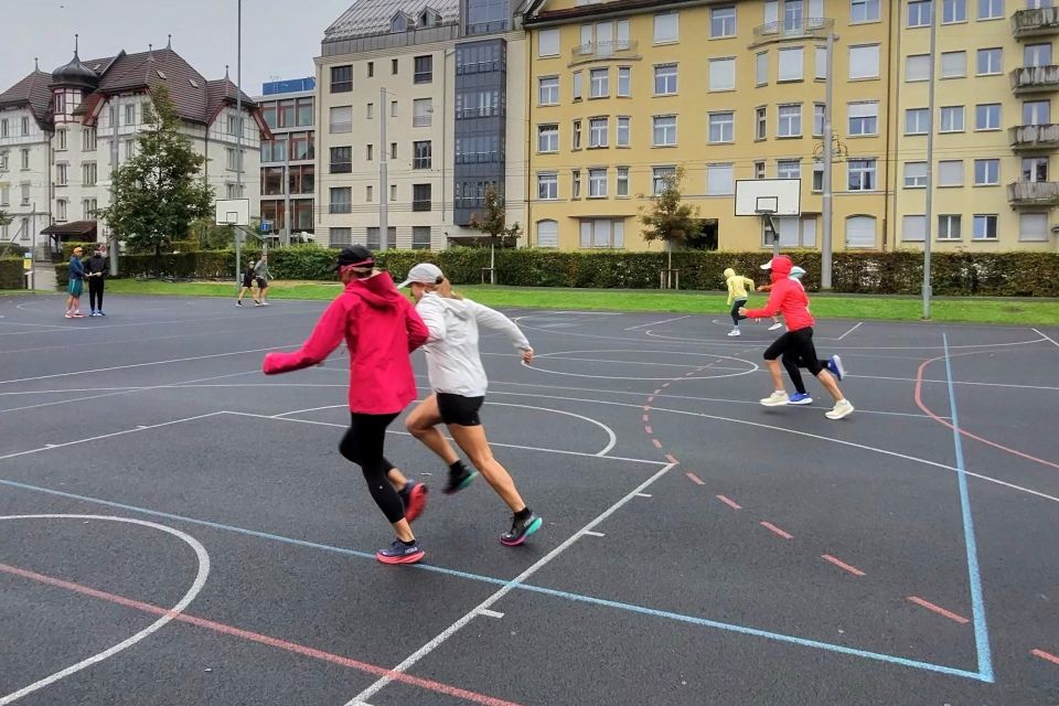 Die meisten Spieler tragen Jacken und Mützen, was auf kühles Wetter hindeutet. Im Hintergrund sind mehrere Gebäude mit vielen Fenstern zu sehen.