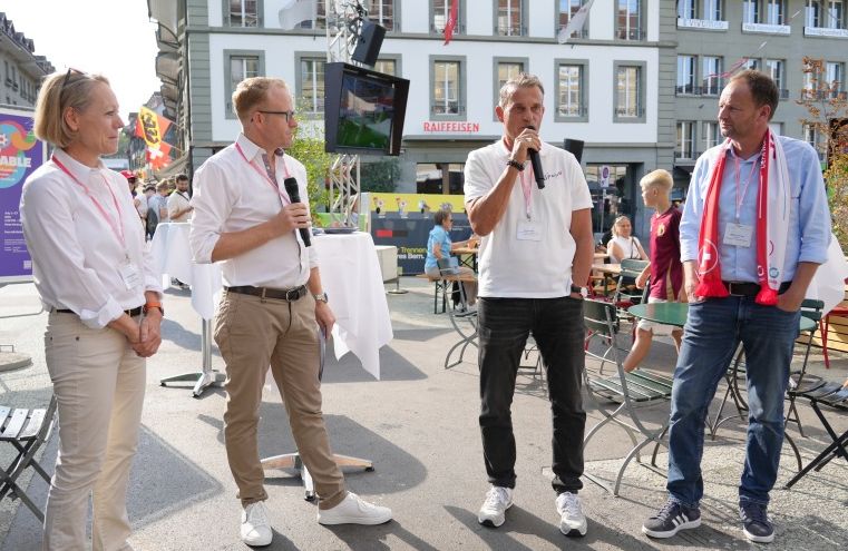 Vier Männer stehen und sprechen mit Mikrofonen bei einer Veranstaltung im Freien auf einem Stadtplatz, umgeben von Menschen, die an Tischen sitzen. Im Hintergrund sind Gebäude, Fahnen und Veranstaltungsdekorationen zu sehen.