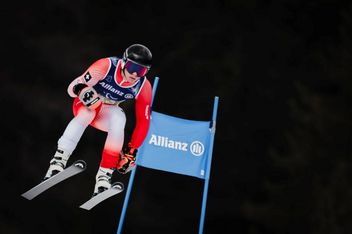 Ein Skispringer in einem rot-weißen Anzug schwebt während eines nächtlichen Wettkampfs mit Helm, Schutzbrille und Handschuhen vor einem dunklen Hintergrund an einer Allianz-Flagge vorbei.