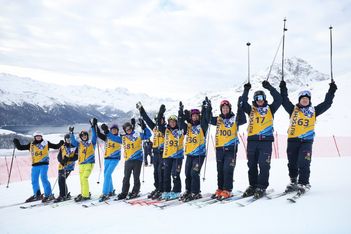 Zehn Skifahrer in farbenfroher Skikleidung und nummerierten Westen stehen in einer Reihe auf einem verschneiten Berg, halten ihre Skistöcke hoch und lächeln, während im Hintergrund schneebedeckte Gipfel und ein See zu sehen sind.